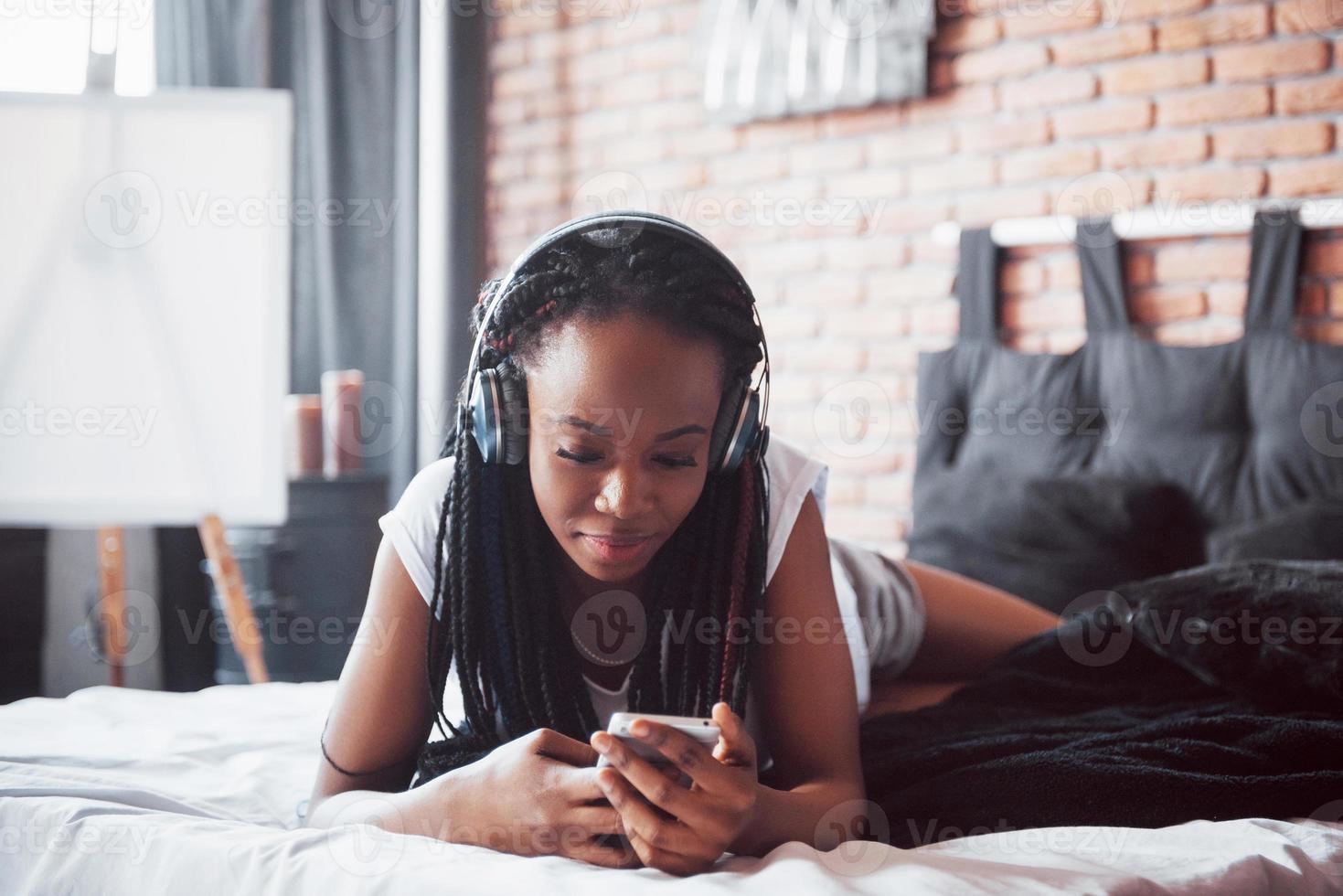bella ragazza africana in pigiameria sorridente ascoltando musica in cuffia sdraiata sul letto foto