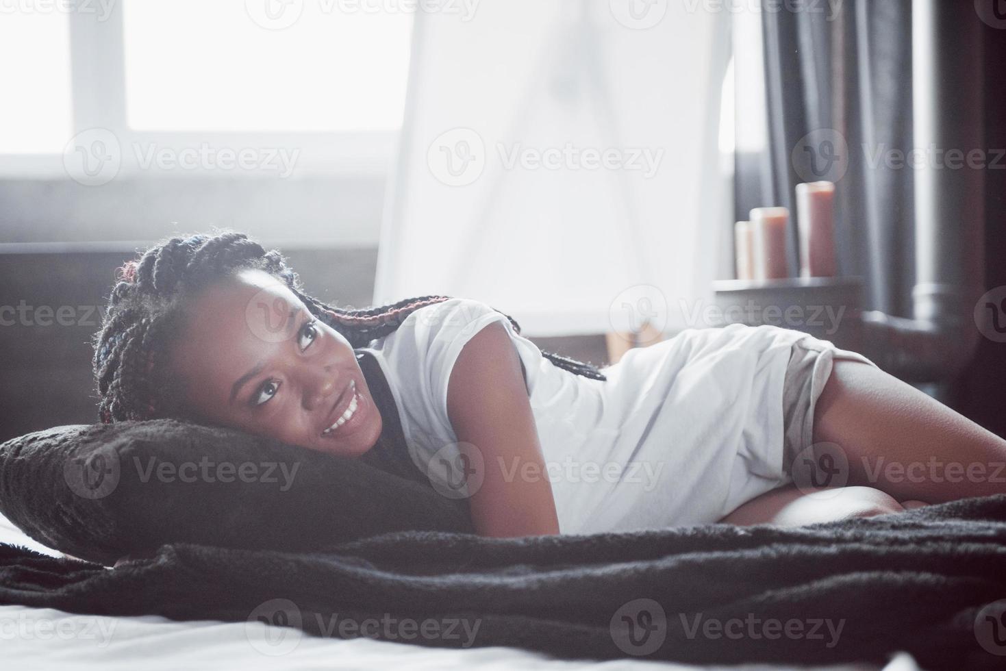 una bella donna africana giace e dorme a letto, in una camera da letto bianca. buongiorno, vacanza a casa. la ragazza indossa una t-shirt, codini in testa foto