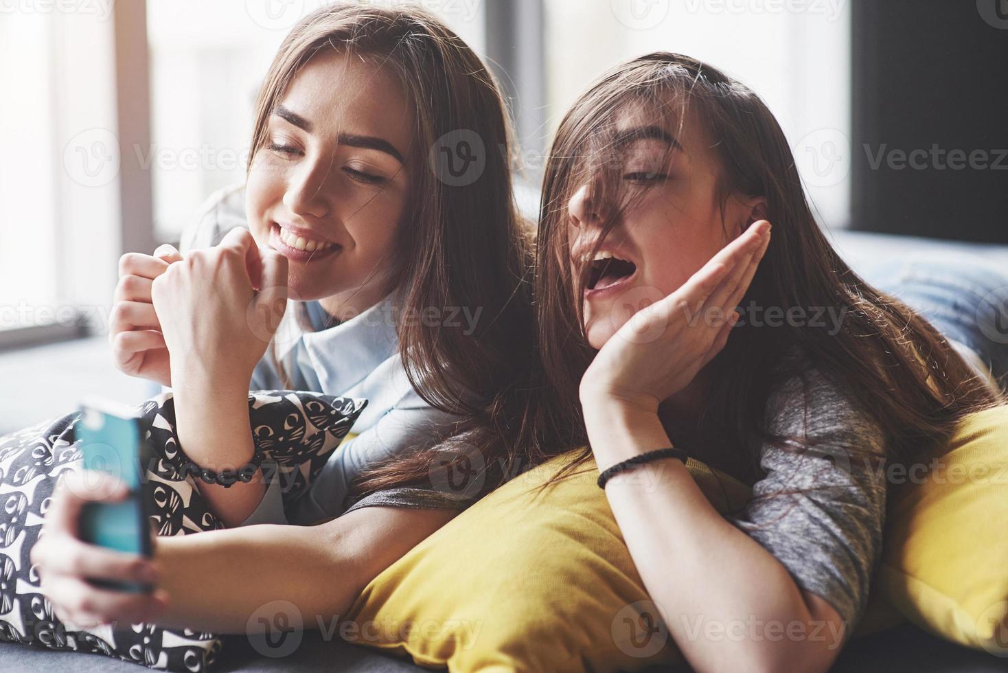 due sorelle gemelle sorridenti carine che tengono smartphone e fanno selfie. le ragazze si sdraiano sul divano in posa e gioia foto