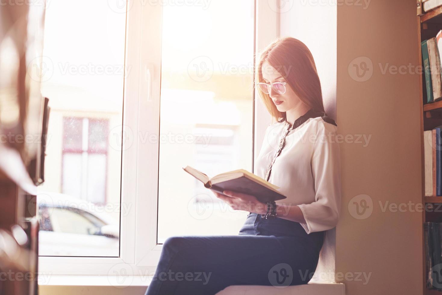 una studentessa si siede sul davanzale di una finestra in una biblioteca e legge un libro foto
