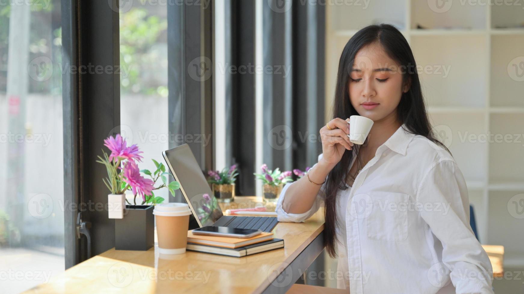 giovane donna che indossa un abito bianco in possesso di un caffè da bere mentre si riposa da un laptop che lavora in un ufficio. foto