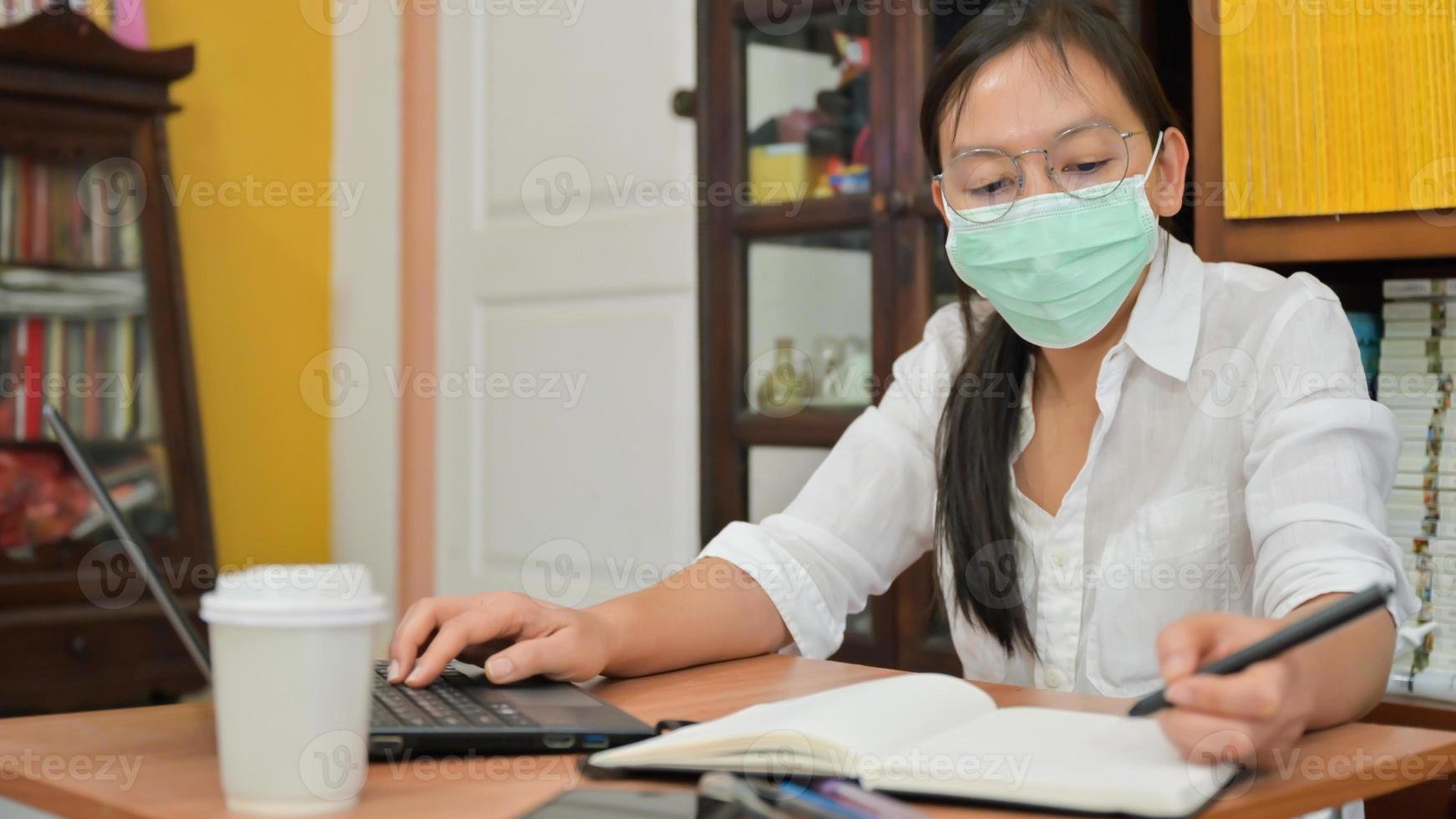 la donna asiatica sta prendendo appunti e sta usando un laptop. lavora a casa per proteggersi dal virus corona o covid-19. foto