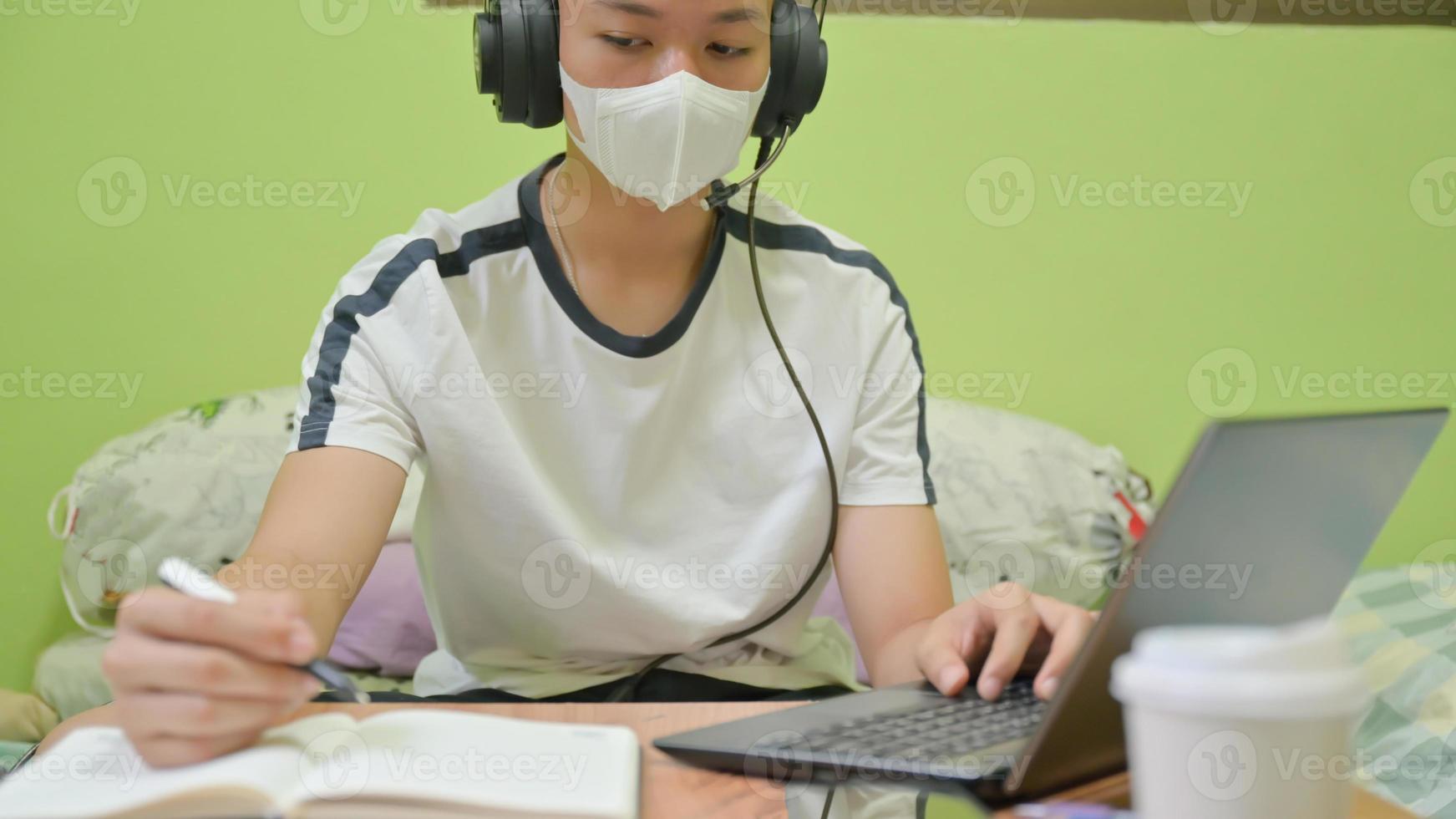 studente maschio con maschera che usa un laptop e prende appunti per la preparazione dell'esame. Ha studiato a casa per proteggere covid-19. foto