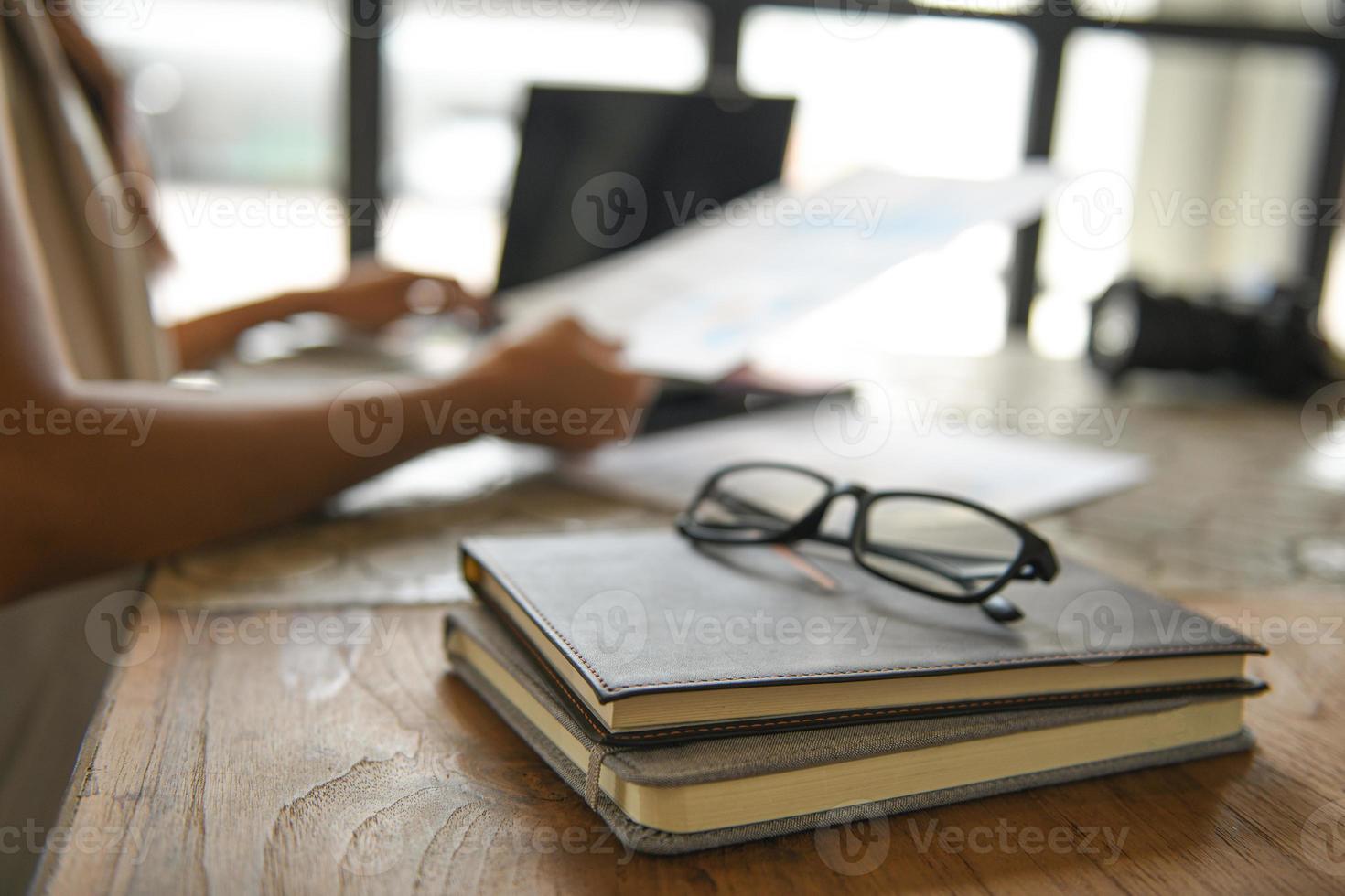 occhiali posizionati su quaderni, sfondo sfocato le persone sono sedute usando il laptop. foto