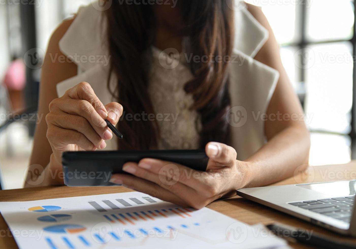 la mano della donna sta usando una penna sullo schermo di un telefono cellulare. grafici e laptop posizionati sulla scrivania. foto