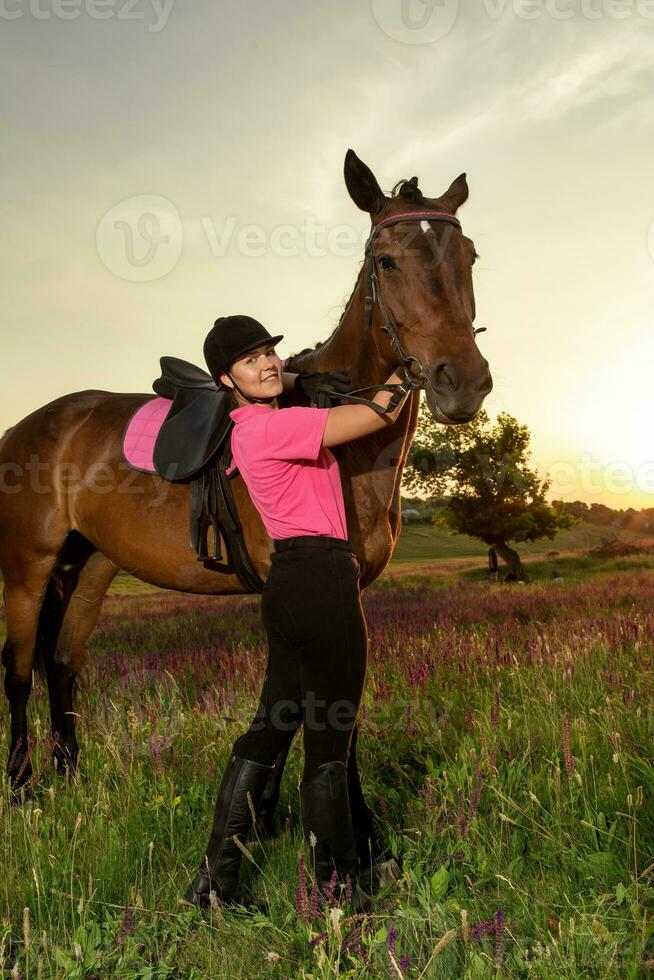 bellissimo giovane ragazza Sorridi a sua cavallo condimento uniforme concorrenza all'aperto ritratto su tramonto foto