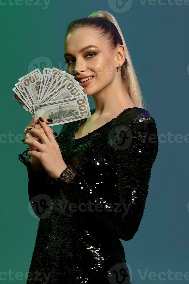 bionda femmina nel gioielleria e nero lustrino vestire. sorridente, mostrando alcuni centinaio dollaro fatture, in posa su colorato sfondo. poker, casinò. avvicinamento foto