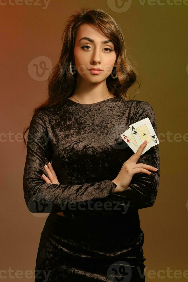brunetta femmina nel nero velluto vestito e gioielleria mostrando Due assi, in posa contro colorato studio sfondo. gioco d'azzardo, poker, casinò. avvicinamento. foto
