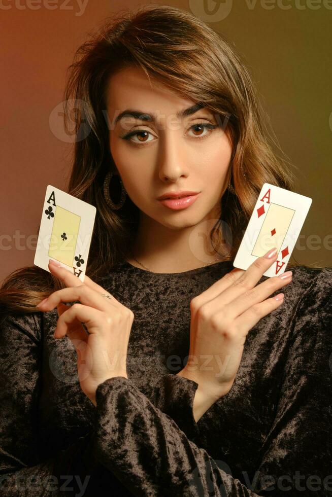 brunetta fanciulla nel nero velluto vestito e gioielleria mostrando Due assi, in posa contro colorato studio sfondo. gioco d'azzardo, poker, casinò. avvicinamento. foto