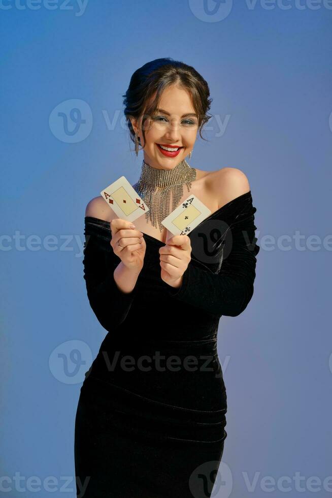 brunetta femmina con spoglio le spalle, nel nero vestito e gioielleria. sorridente, mostrando Due assi, in posa su blu sfondo. poker, casinò. avvicinamento foto