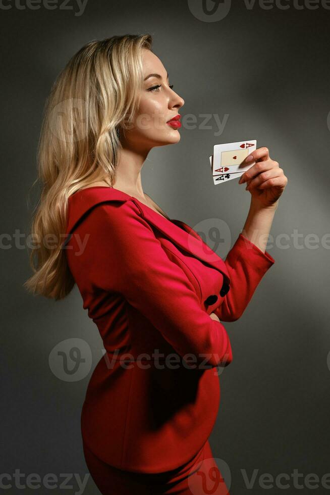 bionda modello nel rosso vestito e nero orecchini. lei mostrando Due giocando carte, in posa lateralmente su grigio studio sfondo. poker, casinò. avvicinamento foto