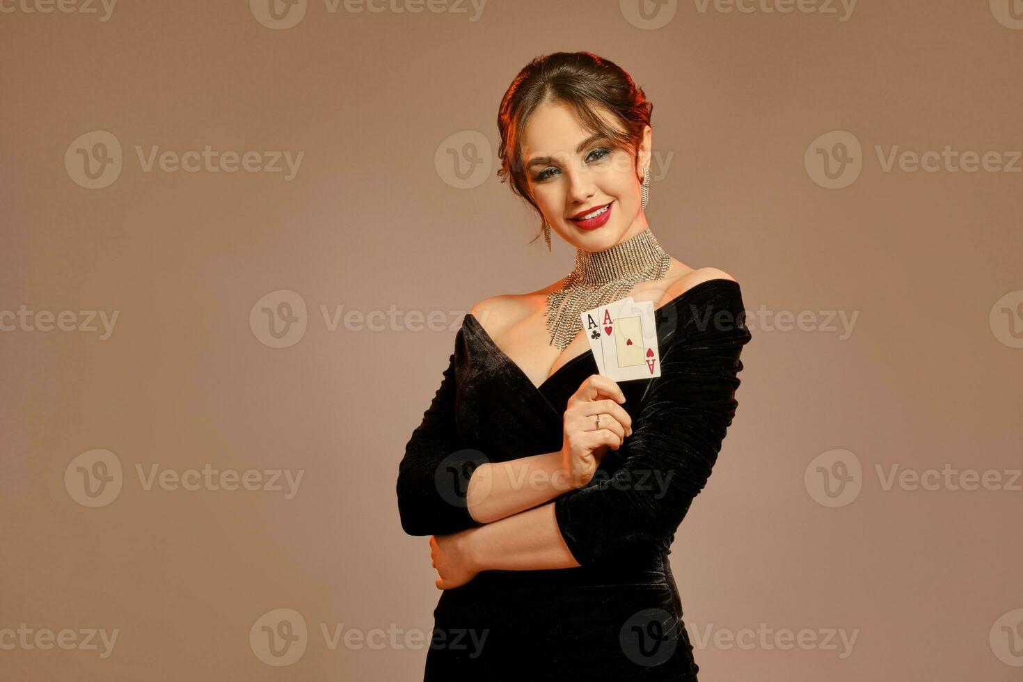 brunetta femmina, spoglio le spalle, nel nero vestito e gioielleria. sorridente, mostrando Due assi, in posa su Marrone studio sfondo. poker, casinò. avvicinamento foto