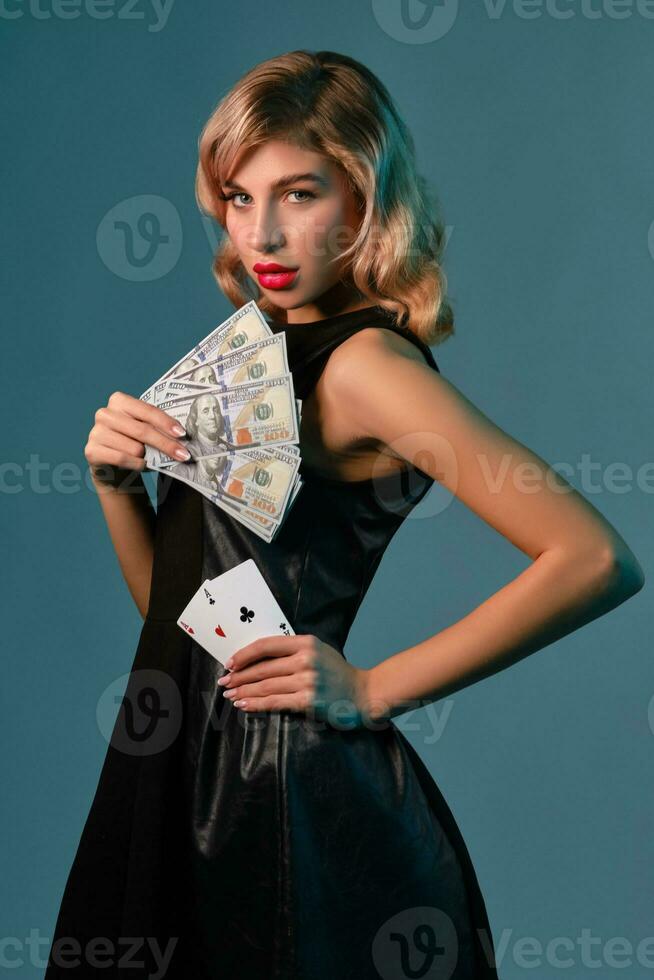 bionda ragazza nel nero elegante vestito Tenere alcuni i soldi e carte, in posa contro blu sfondo. gioco d'azzardo intrattenimento, poker, casinò. avvicinamento. foto