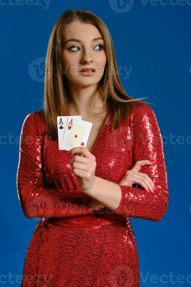 bionda modello con trucco, nel rosso lustrino vestito è mostrando Due assi, mani piegato, in posa su blu sfondo. gioco d'azzardo, poker, casinò. avvicinamento. foto
