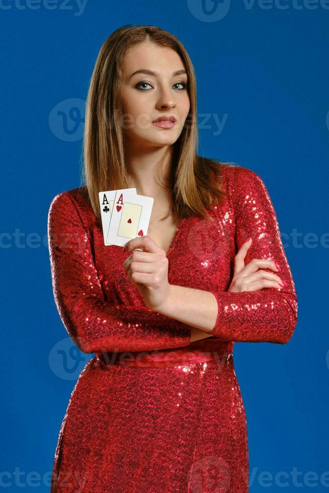 bionda donna con trucco, nel rosso lustrino vestito è mostrando Due assi, mani piegato, in posa su blu sfondo. gioco d'azzardo, poker, casinò. avvicinamento. foto