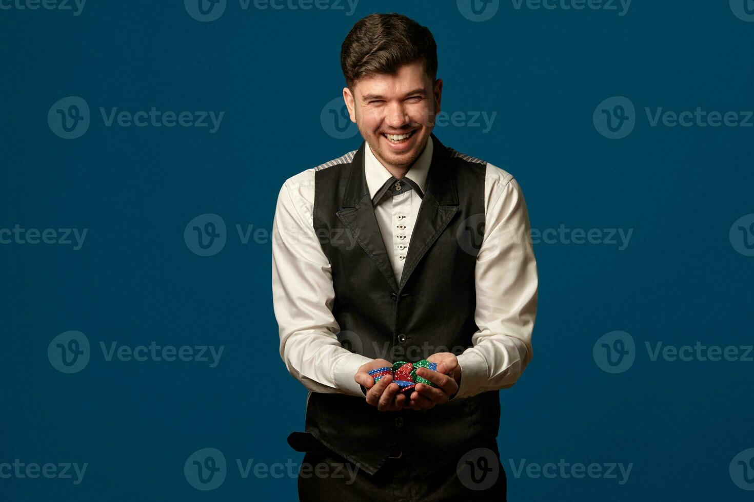 novizio nel poker, nel nero veste e i pantaloni, bianca camicia. Tenere alcuni colorato patatine fritte. in posa contro blu sfondo. gioco d'azzardo, casinò. avvicinamento. foto