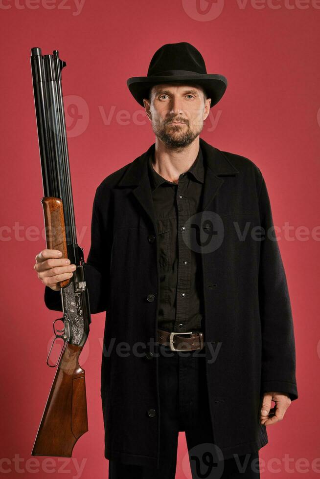 di mezza età uomo con barba, baffi, nel nero giacca e cappello, Tenere un' pistola mentre in posa contro un' rosso sfondo. sincero emozioni concetto. foto