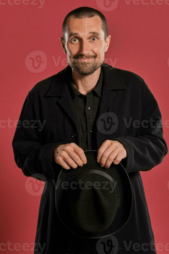 di mezza età uomo con barba e baffi, indossa nero cappello e giacca in posa contro un' rosso sfondo. sincero emozioni concetto. foto