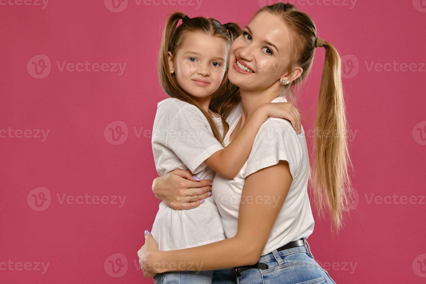 mamma e figlia con un' divertente code di cavallo, vestito nel bianca magliette e blu denim jeans siamo in posa contro un' rosa studio sfondo. avvicinamento sparo. foto