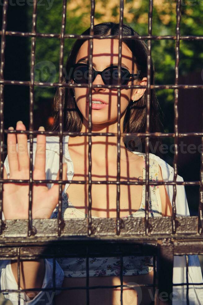 ritratto di ragazza nel occhiali da sole in posa nel città dietro a un' a traliccio recinzione. vestito nel superiore con floreale Stampa, bianca camicia, nero i pantaloni, vita Borsa. foto