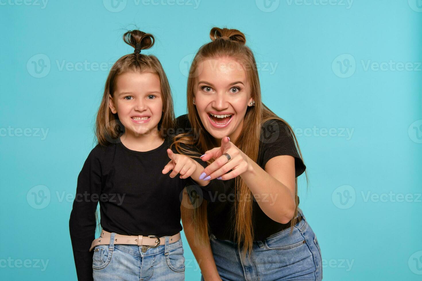 mamma e figlia con un' divertente acconciature, vestito nel nero camicie e blu denim jeans siamo in posa contro un' blu studio sfondo. avvicinamento sparo. foto