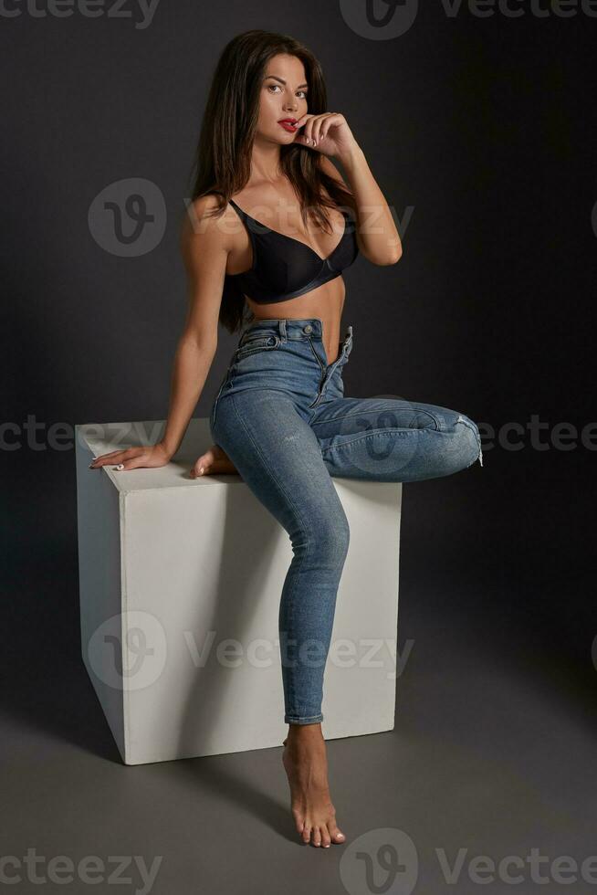 bellissimo sexy brunetta donna in posa nel studio, seduta, guardare a telecamera. ragazza indossare alla moda jeans e sensuale biancheria intima. foto