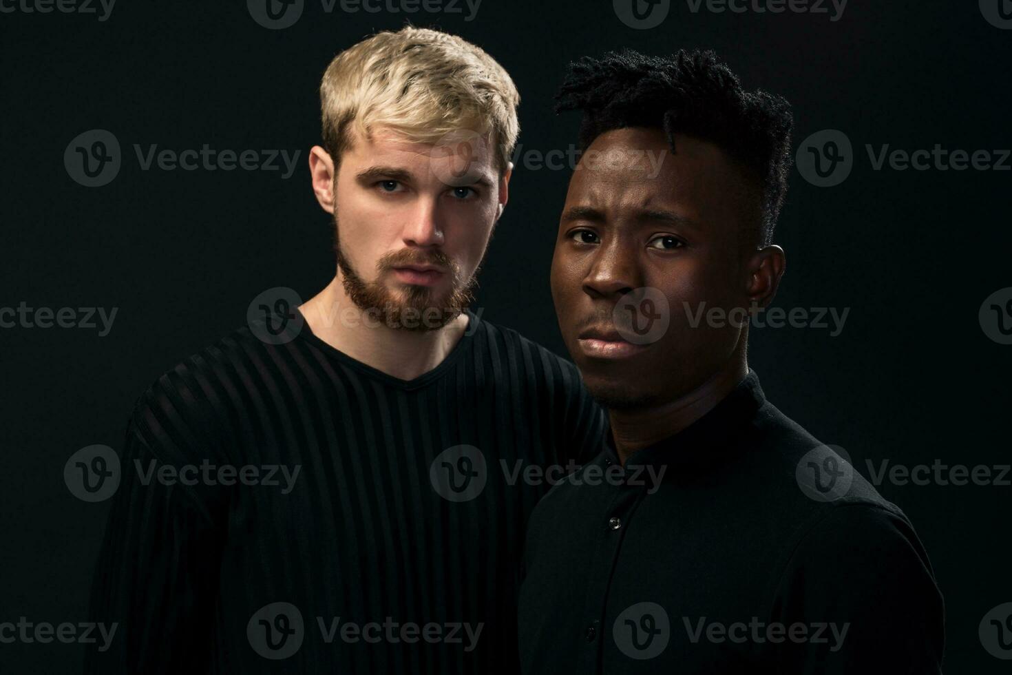 ritratto di Due giovane africano americano e caucasico uomini in piedi al di sopra di nero sfondo. studio tiro foto