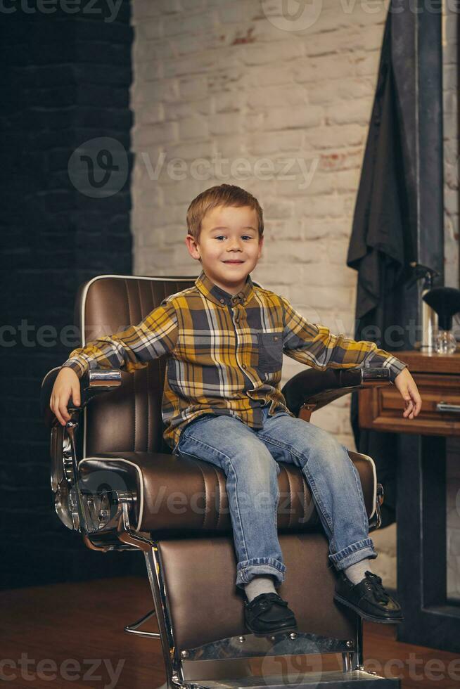 ritratto di un' elegante poco ragazzo vestito nel camicia e jeans nel il barbiere, seduta nel un' sedia contro il del barbiere posto di lavoro foto