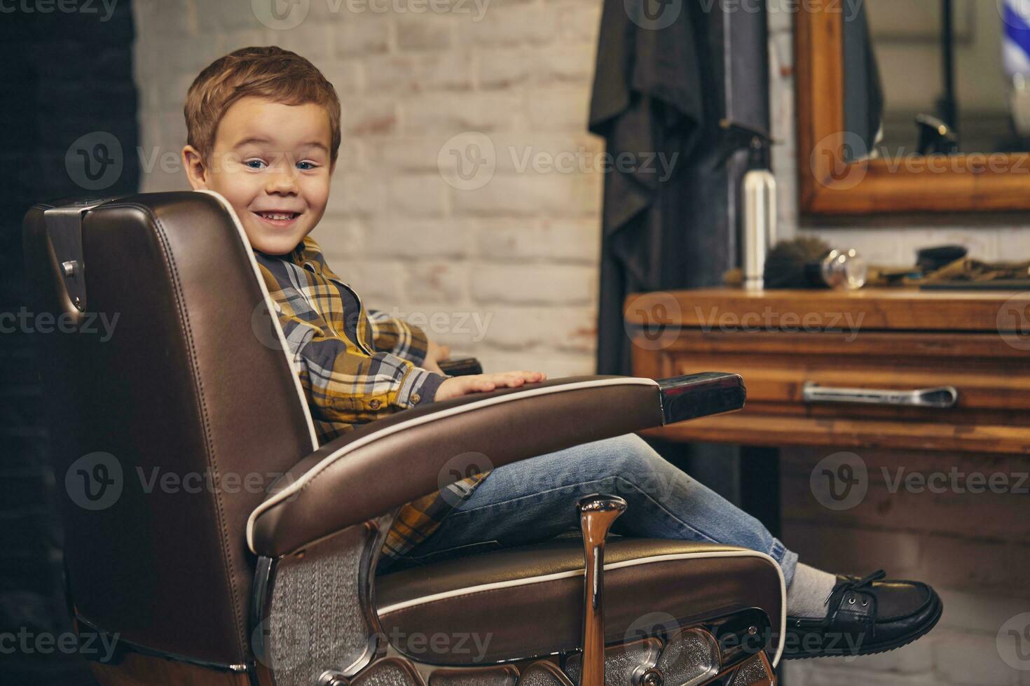 ritratto di un' elegante poco ragazzo vestito nel camicia e jeans nel il barbiere, seduta nel un' sedia contro il del barbiere posto di lavoro foto