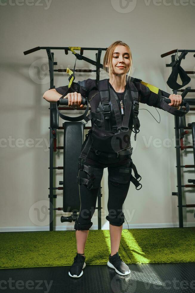 bellissimo bionda donna indossare nel sensori e cinghie, si allaccia con velcro è praticante ems fitness nel un' Palestra l'esecuzione trazione esercizi. foto