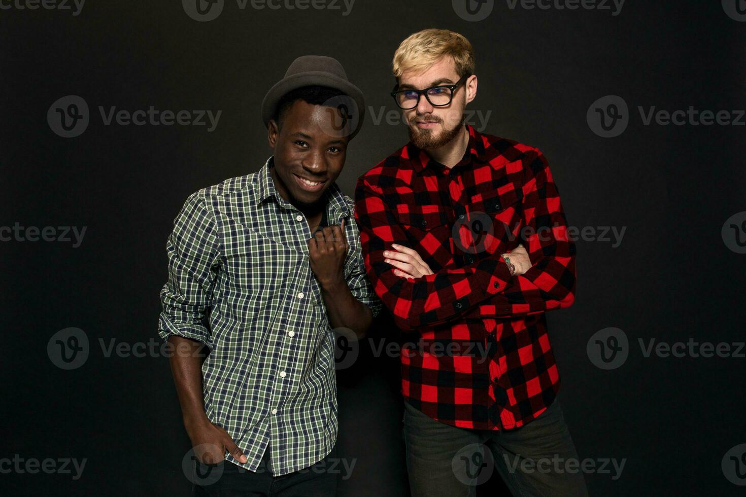 studio tiro di Due elegante giovane uomini avendo divertimento. bello barbuto fricchettone nel un' camicia nel un' gabbia in piedi Il prossimo per il suo afroamericano amico nel cappello contro un' buio sfondo. foto