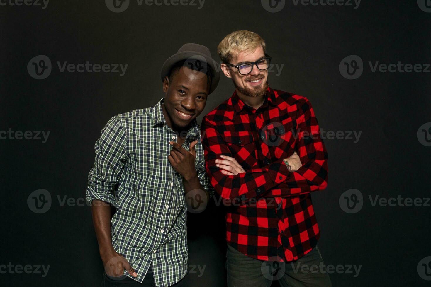 studio tiro di Due elegante giovane uomini avendo divertimento. bello barbuto fricchettone nel un' camicia nel un' gabbia in piedi Il prossimo per il suo afroamericano amico nel cappello contro un' buio sfondo. foto