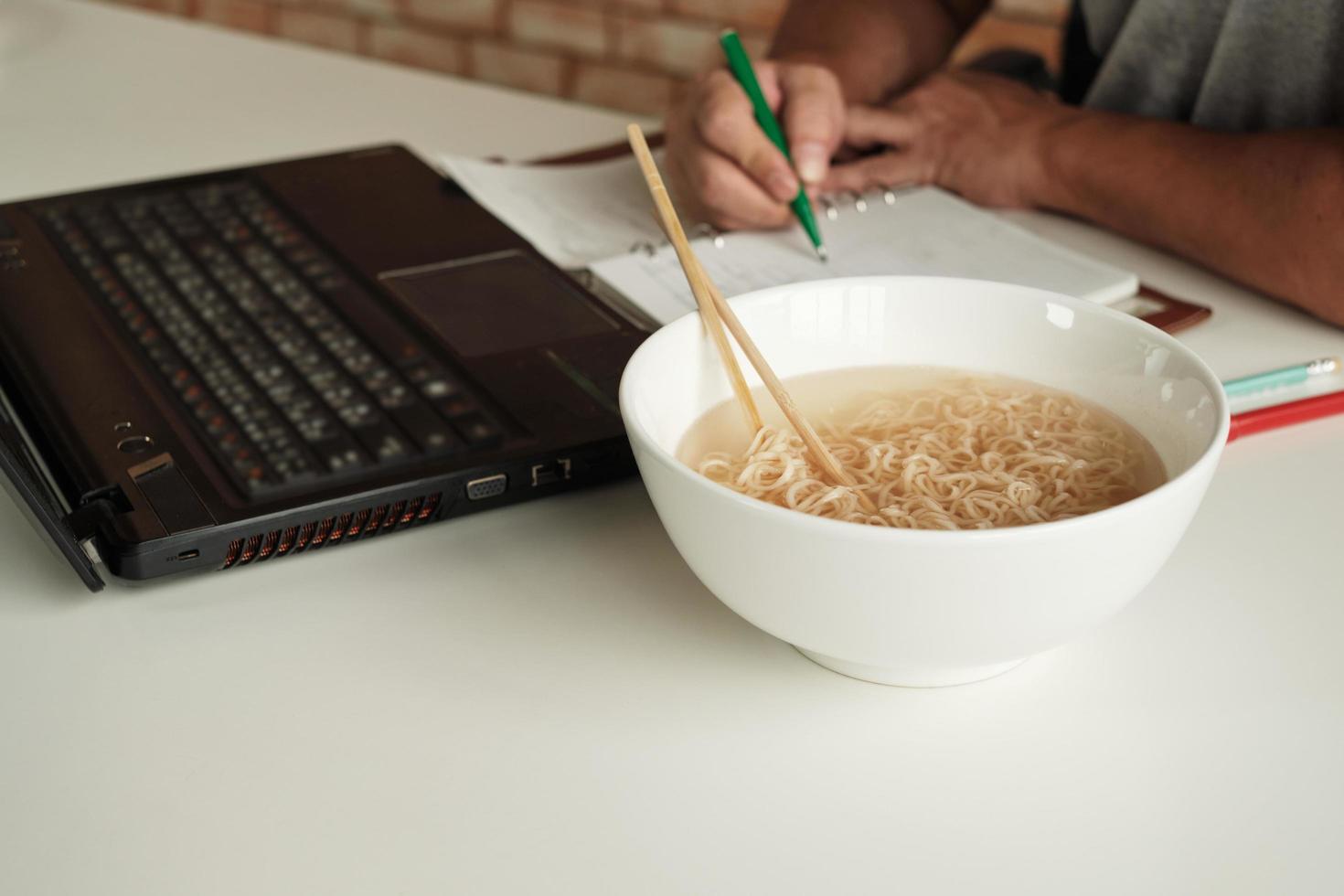 lavoratore di sesso maschile tailandese impegnato a lavorare con il laptop, usa le bacchette per mangiare frettolosamente noodles istantanei durante la pausa pranzo in ufficio, perché veloce, gustoso ed economico. nel tempo fast food asiatico, stile di vita malsano. foto