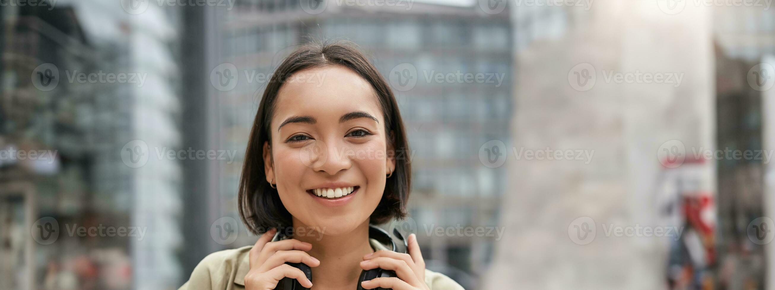 verticale tiro di bellissimo asiatico donna in posa con cuffie in giro collo, sorridente e ridendo, in piedi su strada nel luce del giorno foto