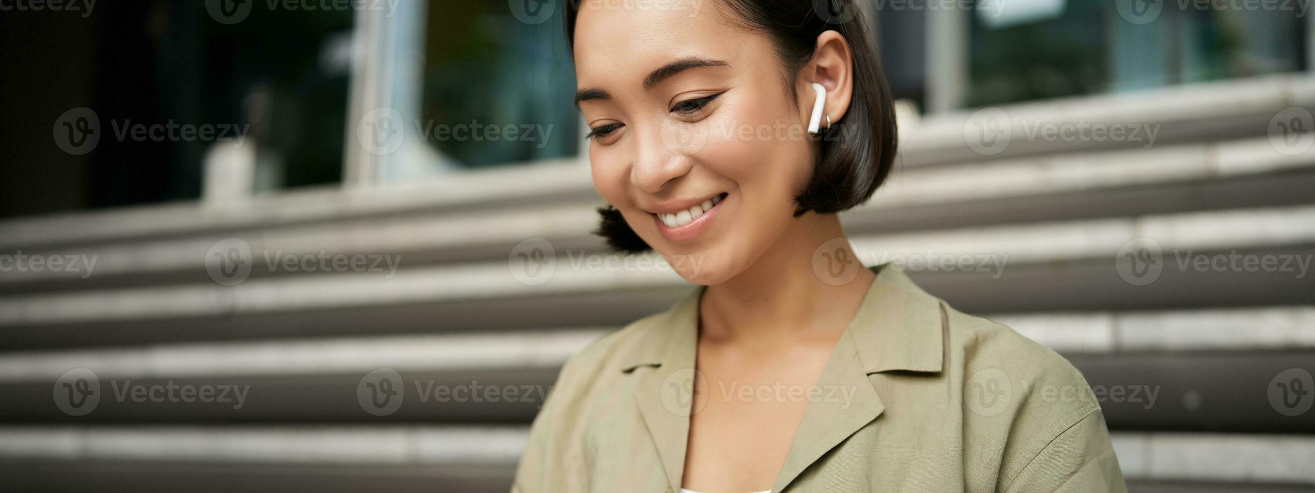 ritratto di sorridente asiatico ragazza ascolta musica, podasta nel senza fili auricolari, utilizzando cuffie all'aperto, seduta su strada foto