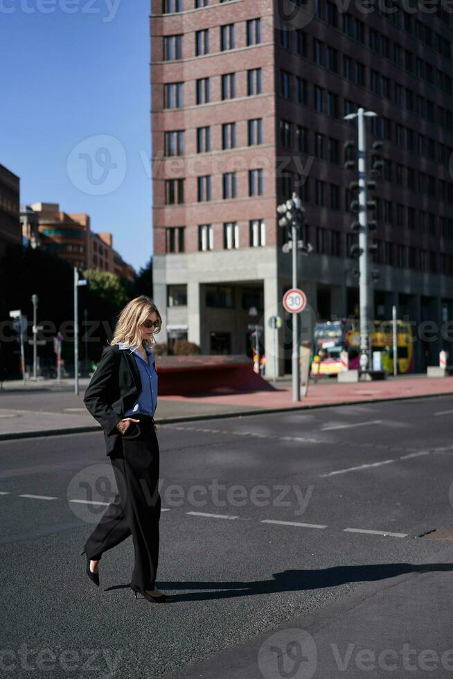 verticale ritratto di fiducioso e elegante donna d'affari nel completo da uomo, a passeggio, attraversare il strada nel città centro, indossa occhiali da sole foto