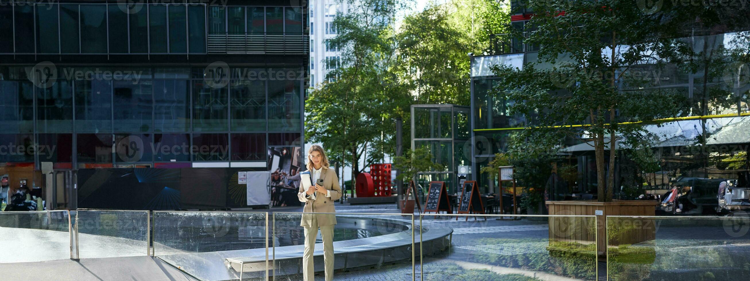 aziendale donna nel completo da uomo, in piedi con fiducia nel beige completo da uomo con Lavorando documenti all'aperto, in posa nel città centro foto