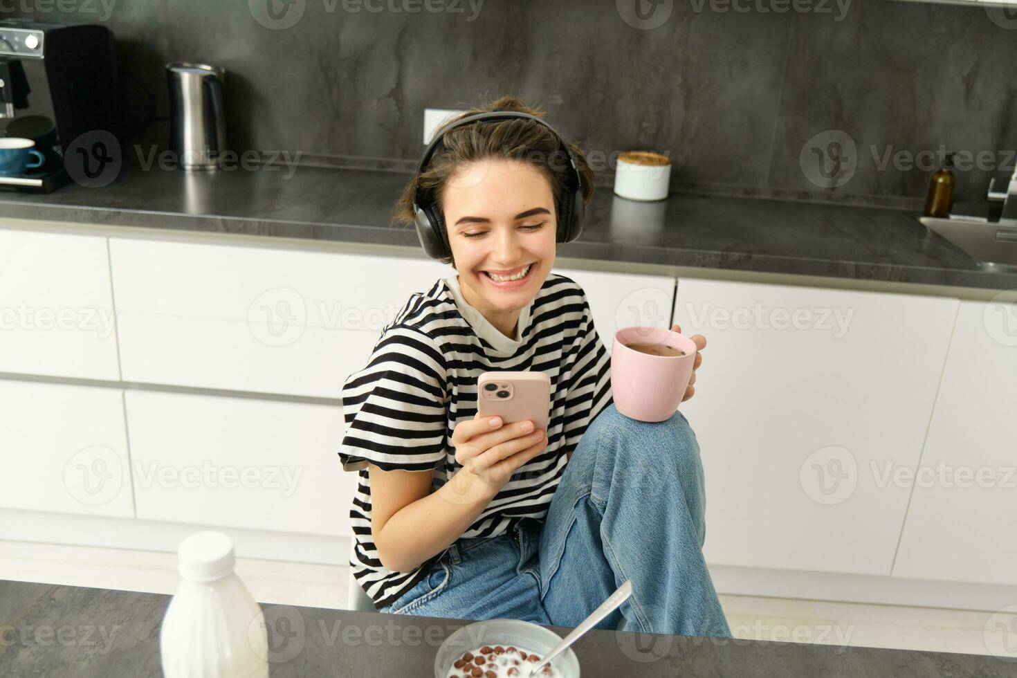 moderno femmina modello sorridente, utilizzando mobile Telefono, potabile tè e mangiare prima colazione nel cucina foto