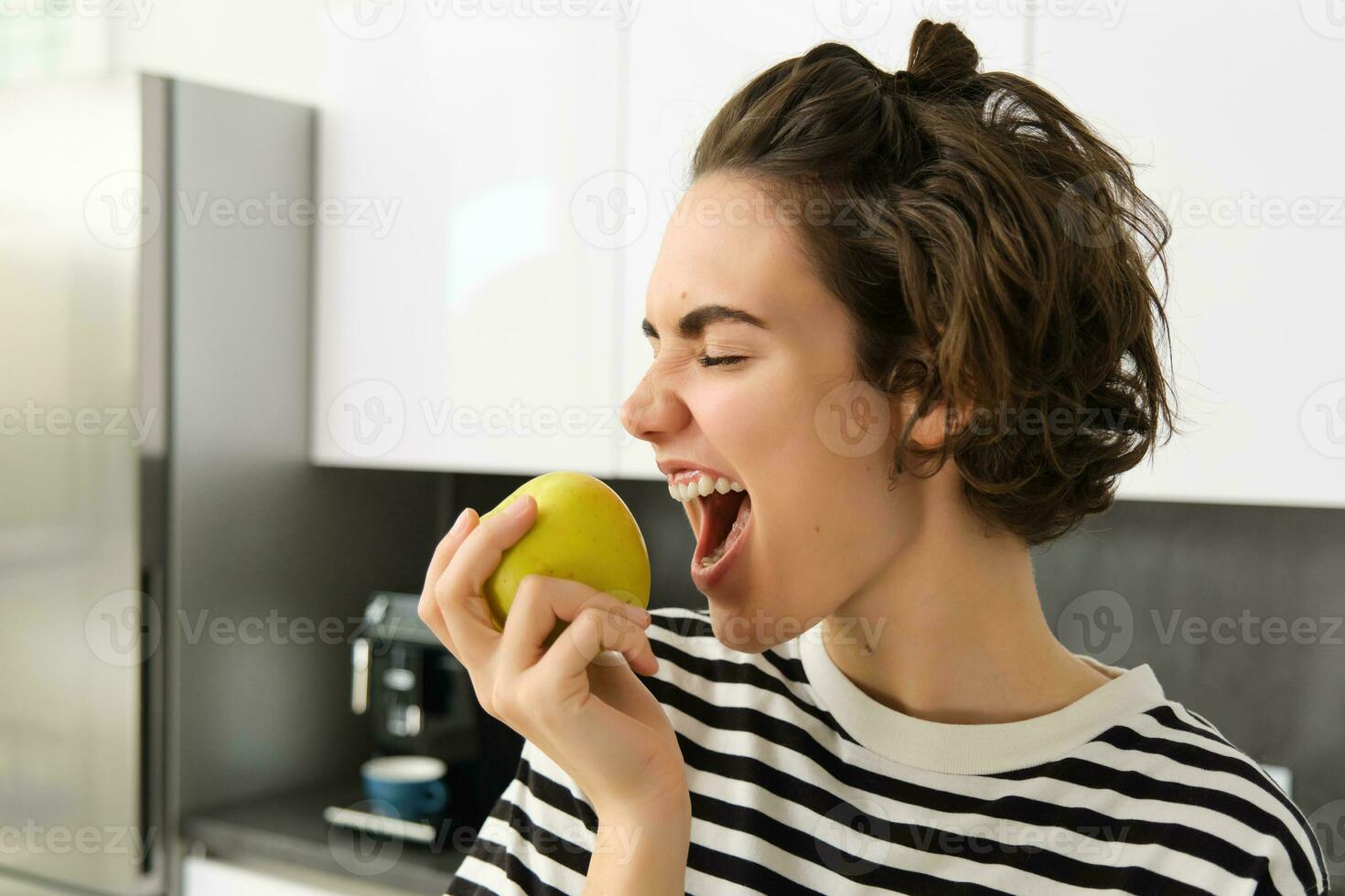 vicino su ritratto di sorridente donna nel il cucina, Tenere un mela, mangiare frutta, avendo un' salutare merenda per pranzo a casa foto