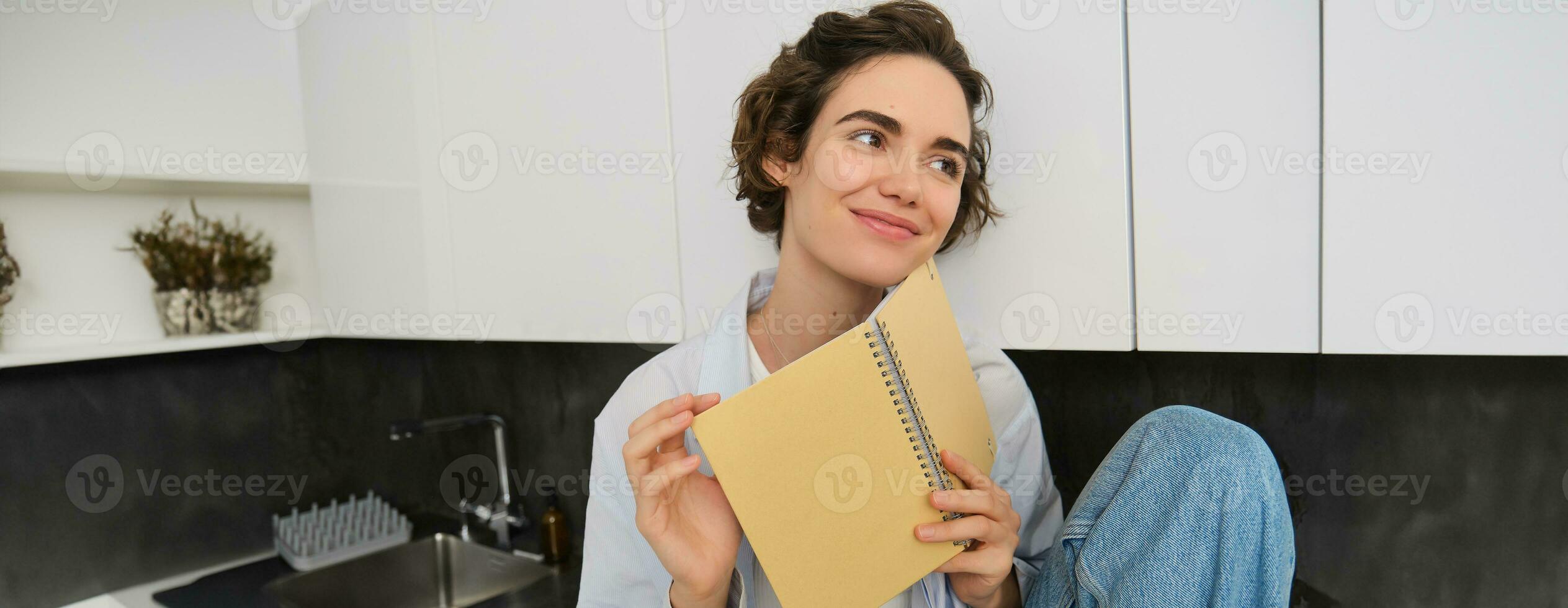 ritratto di giovane moderno donna seduta nel cucina, capovolgimento pagine, lettura rivista prenotare, sorridente felicemente foto