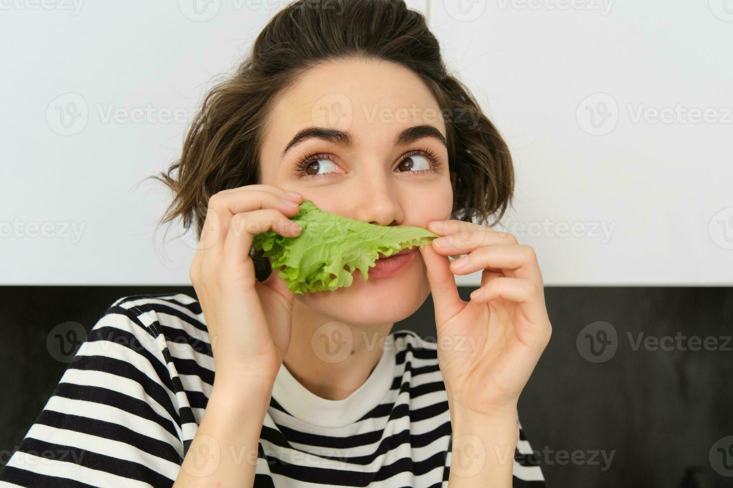 vicino su di divertente carino donna, vegetariano mangiare lattuga foglia e sorridente, concetto di salutare dieta, ragazza piace la verdura, in piedi nel il cucina foto