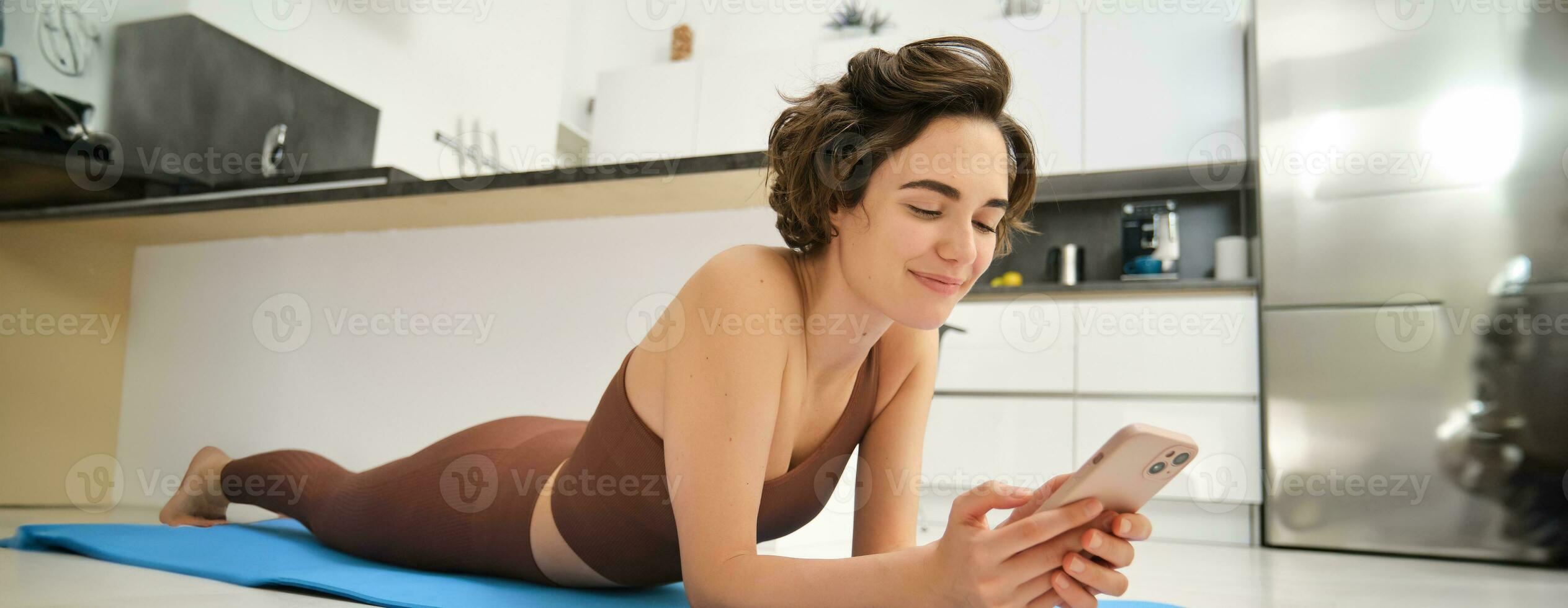 Immagine di sportivo ragazza riposo su yoga gomma da cancellare stuoia, allenarsi a casa, utilizzando sua smartphone, guardare per in linea fitness tutorial, mobile App per formazione foto