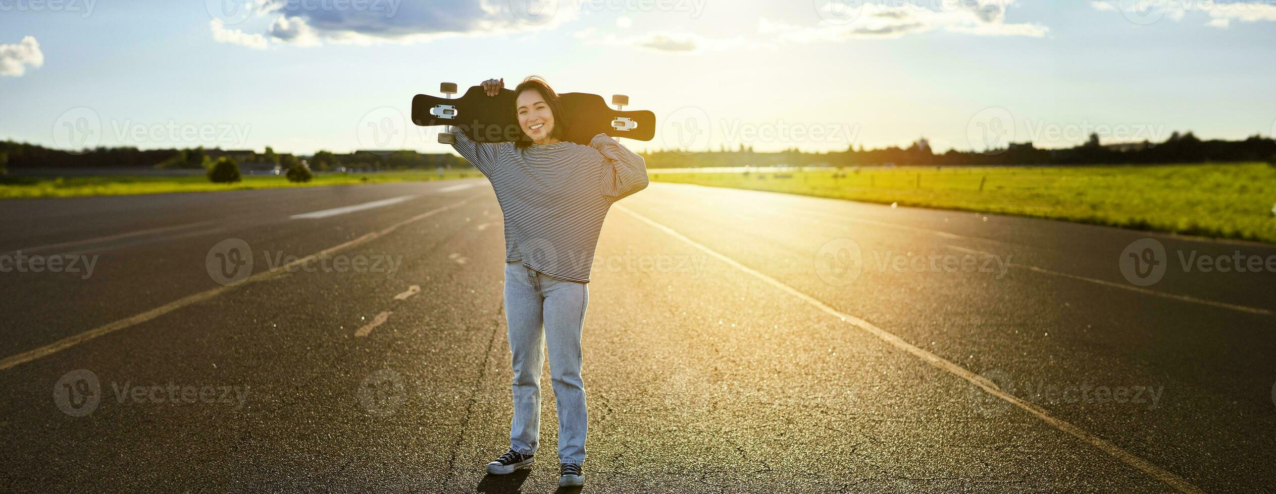 giovane asiatico donna in piedi con longboard su soleggiato strada, pattinando nel pattinare parco su sua incrociatore foto