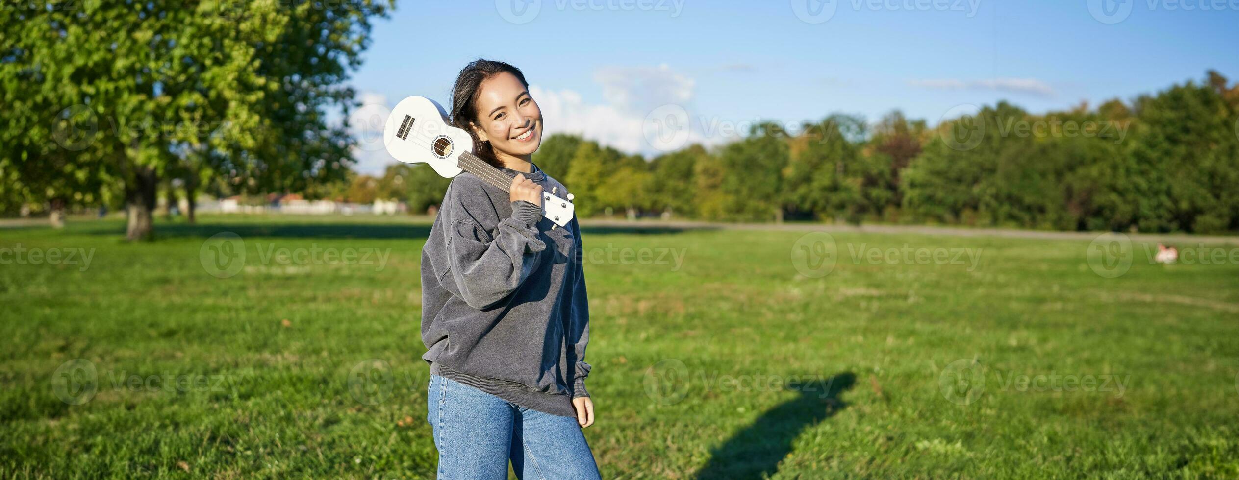 bellissimo coreano ragazza in posa con ukulele, giovane musicista giocando all'aperto nel verde parco su soleggiato giorno foto