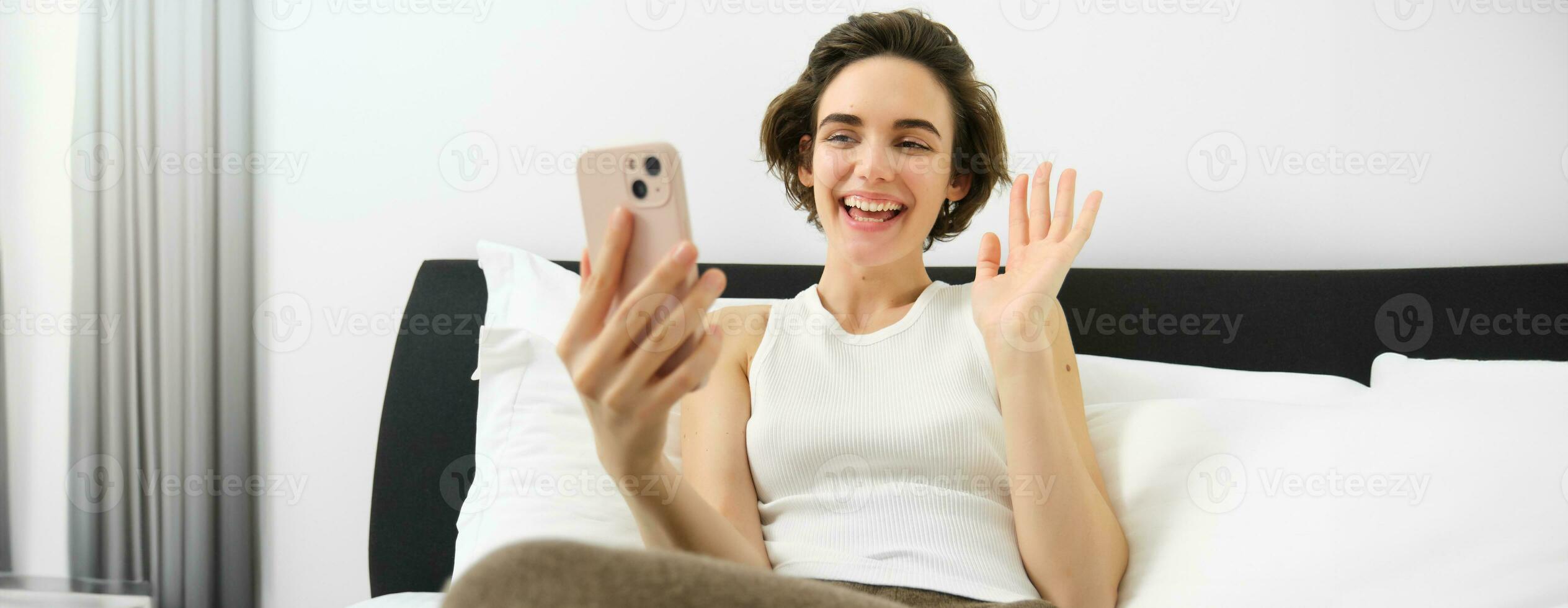 allegro sorridente donna riposo nel letto, si connette per in linea video chiamata mentre dire bugie nel Camera da letto, parlando per amico attraverso smartphone App foto