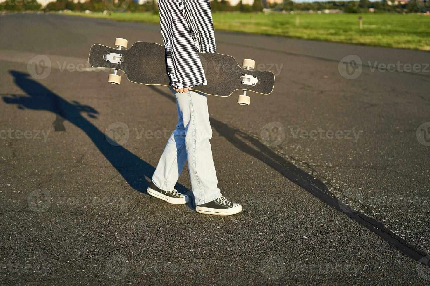 ritagliata tiro di adolescente ragazza corpo, Tenere incrociatore longboard nel mano, a piedi nel scarpe da ginnastica su strada nel jeans e felpa. giovane donna pattinatore con skateboard foto