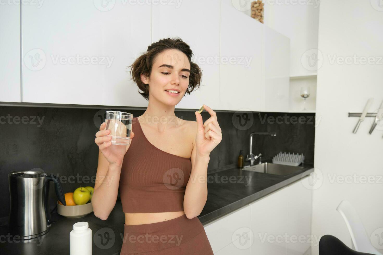dietetico integratori e salutare stile di vita. giovane donna assunzione vitamina c, d Omega 3 con bicchiere di acqua, in piedi nel abbigliamento sportivo, potabile dopo allenarsi formazione nel sua cucina foto