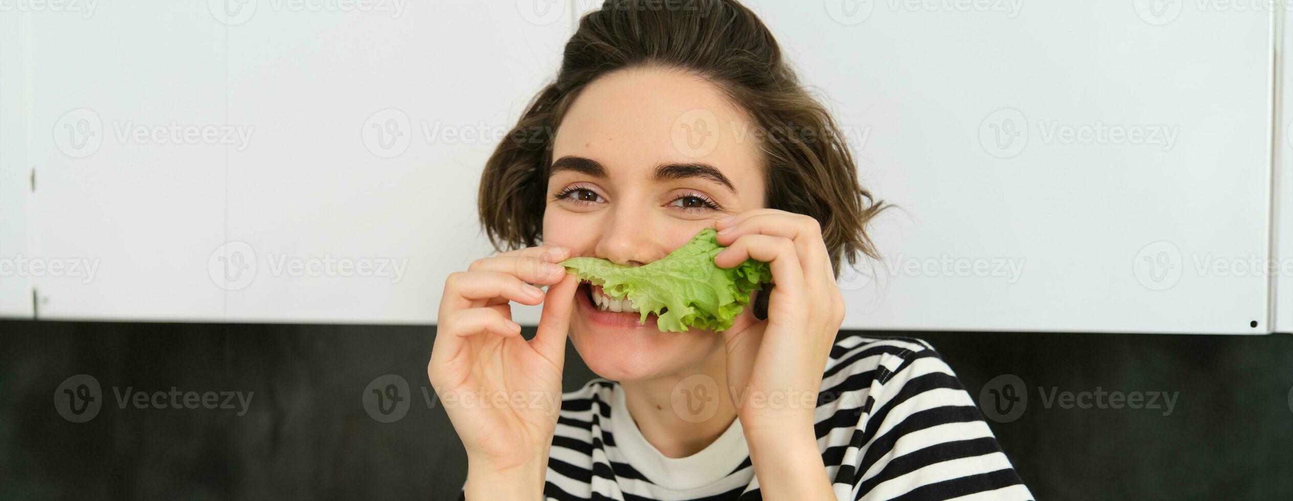 vicino su ritratto di giovane donna, vegetariano ragazza, piace mangiare la verdura, in posa con lattuga foglia e sorridente, in posa nel il cucina. concetto di salutare cibo e dieta foto