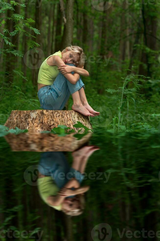 adolescenziale ragazza si siede su un' ceppo nel un' naturale foresta parco con un' triste e riflessivo Guarda, riflessa nel il acqua foto