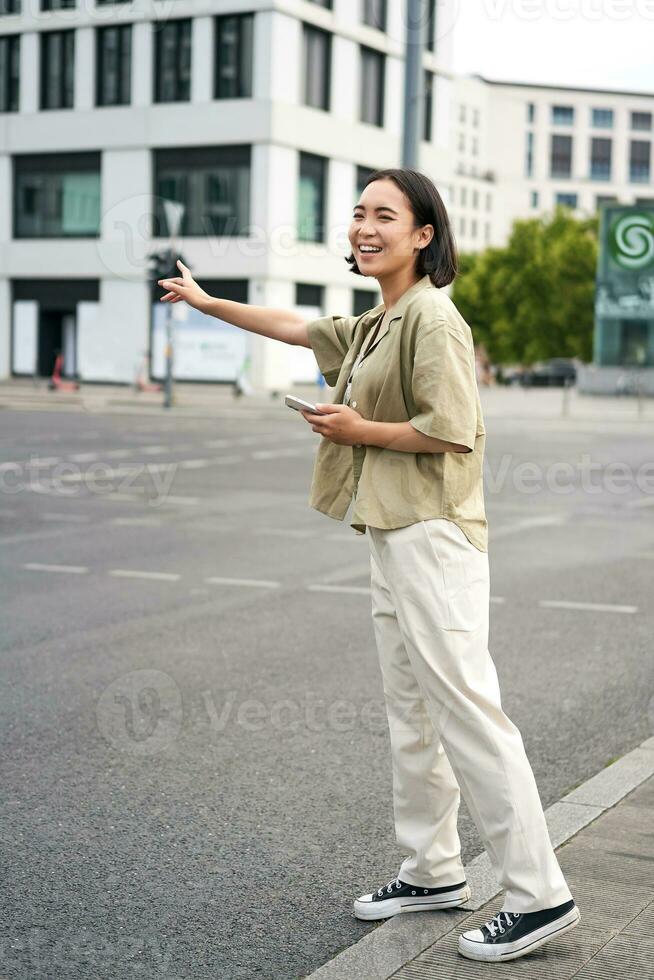 verticale tiro di giovane donna provando catturare Taxi, agitando a autista su strada, Tenere smartphone con auto condivisione app, in piedi su città strada foto